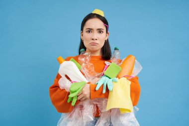 Elinde boş plastik şişeler ve çöplerle, geri dönüşüm için atık ayıklayan kızgın genç bir kadın, ellerinde mavi arka planda, düzensiz bir çöpten dolayı üzgün görünüyor..