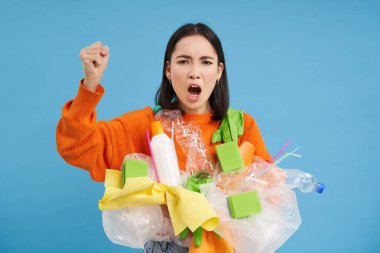 Kızgın Koreli kadın bağırıyor, plastik atıklar tutuyor, çöpleri geri dönüştürmeye teşvik ediyor, yumruk sallıyor, mavi arka plan.