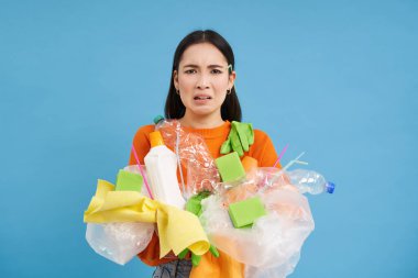 Elinde plastik çöplerle suratını asan Asyalı kadın, geri dönüşüm atığından şikayet ediyor, mavi arka planda çöplerle dikiliyor..
