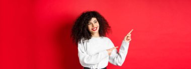 Kıvırcık saçlı ve makyajlı güzel bir Avrupalı kadın, parmaklarını sola uzatıyor ve rüya gibi bir gülümsemeyle yan yana bakıyor, terfi anlaşmasına bakıyor, kırmızı arka plan..