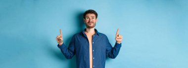 Şüpheci genç adam sırıtıyor ve şüpheli bakıyor, üzgün parmaklarını gösteriyor, mavi arka planda duruyor. Boşluğu kopyala