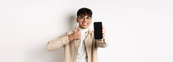 戴着眼镜的英俊年轻人的画像 展示空的智能手机屏幕和大拇指 推荐网络应用或商店 站在白色背景下高兴 — 图库照片