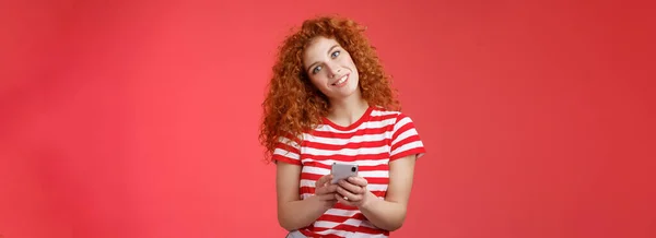 愚かなかわいい赤毛巻き毛のファッショナブルな女性ホールドスマートフォンの傾き頭笑顔広くおもちゃの正の笑顔疑問に思う答えは挑発的なお尻のメッセージを送信赤の背景 — ストック写真