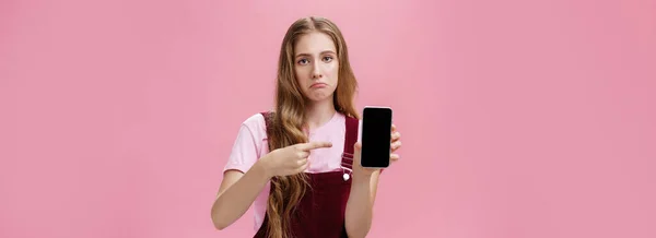 stock image Lifestyle. Sad gloomy young female with cute wavy natural hairstyle showing smartphone screen pointing at gadget with index finger making upset face frowning displeased feeling regret after buying