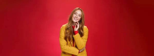 无忧无虑 快乐的红头发女友 穿着黄色的外套 面带微笑 低着头 有着积极的情绪 在红色的背景下显得很开心 — 图库照片