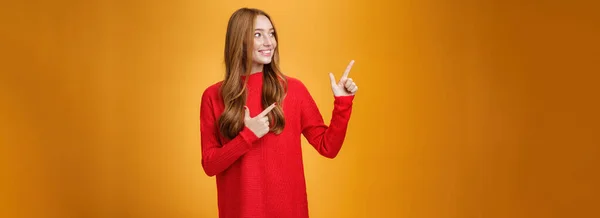 赤暖かいセーターを指して 広い笑顔で左上隅を見て オレンジの背景に素敵な幸せなシーンを楽しんで優しいと柔らかい若い20代の生姜の女性に興味を持っています 広告コンセプト — ストック写真