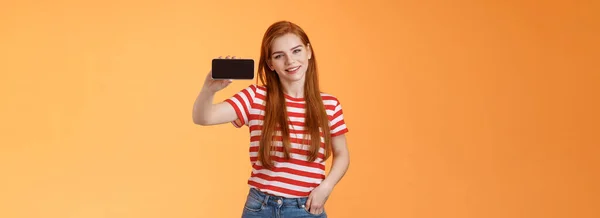 有自信迷人的20多岁的白人姜姑娘介绍智能手机应用 推荐酷游戏 展示手机屏幕水平 微笑自豪 吹牛打分 站橙色背景 — 图库照片