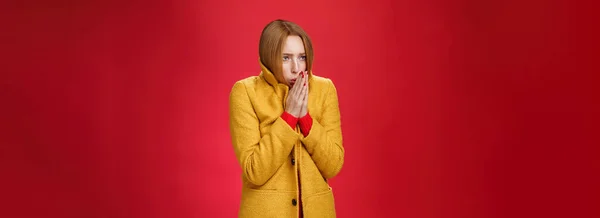 黄色の光のコートを身に着けている強烈な震えかわいい赤毛の女性のスタジオショット手をこすり 暖かい息を手のひらで吹いて 暖かい感じの寒さ 赤の背景に冷たい風の天気の間に凍結 — ストック写真
