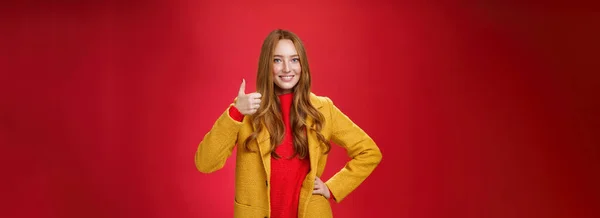 喜びと満足した女性の赤毛の顧客のように親指を表示し 赤の背景に立って新しい黄色のコートの質の高さに満足し 広く幸せな笑みを浮かべて — ストック写真