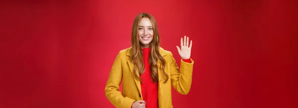 よろしくお願いします フレンドリーなルックスの女性的でスタイリッシュな若いかわいい赤毛の女性黄色の暖かい秋のコートで手を振って挨拶や赤壁の上に広く笑みを浮かべてハイテクジェスチャー — ストック写真