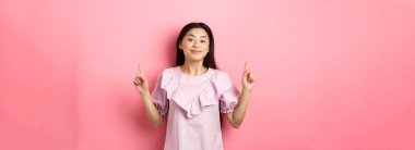 Romantik elbiseli genç Asyalı güzel bir kadın parmaklarını havaya kaldırarak gülümsüyor ve memnuniyetle reklamını yapıyor, pembe arka planda duruyor..