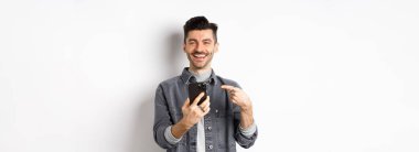 Bıyıklı yakışıklı beyaz adam akıllı telefonu işaret ediyor, gülümsüyor ve cep telefonuyla iyi bir anlaşma yapıyor, beyaz arka plana karşı mutlu duruyor..