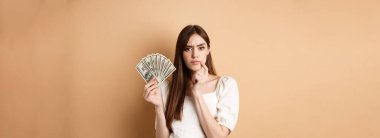 Ciddi ciddi para tutan ve düşünen, kameraya düşünceli görünen, kaşlarını çatan, dolar banknotlarıyla ne alacağına karar veren, bej arka plan.