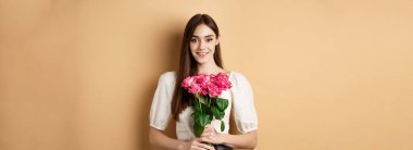 Romantik Sevgililer Günü konsepti. Güzel genç bayan pembe güller tutuyor ve gülümsüyor, bej arka planda mutlu duruyor..