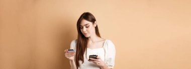 Çevrimiçi ödeme yapan gülümseyen genç kadın, kredi kartına bakıyor ve cep telefonundan alışveriş yapıyor, internetten alışveriş yapıyor, bej arka planda duruyor..