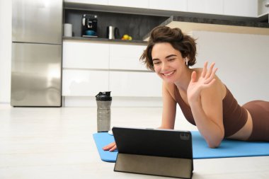 Gülümseyen esmer kadın spor giyim, el sallıyor, tabletten selam veriyor, video spor blogunu kaydediyor, fitness antrenmanını internetten yapıyor, eğitmen egzersiz unsurlarını gösteriyor, mutfakta lastik paspas kullanıyor..