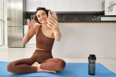 Genç fitness kadınının portresi, evde lastik paspasın üzerinde oturuyor, akıllı telefondan video kaydediyor, vlog yapıyor, evden canlı yayın yapıyor. Boşluğu kopyala