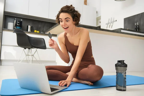 Image of young fitness girl, female athlete at home, workout on kitchen floor, holding credit card with laptop, purchasing online training session, gym application for home workout.