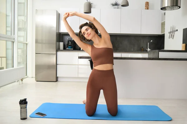 Home training. Young woman workout at home, stretching her body, doing fitness exercises on yoga rubber mat, listening to gym app instructions on smartphone.