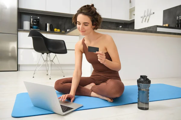 stock image Portrait of young female athlete, yoga girl paying for online classes, purchasing remote training with gym instructor, sitting on her rubber mat with laptop and credit card.