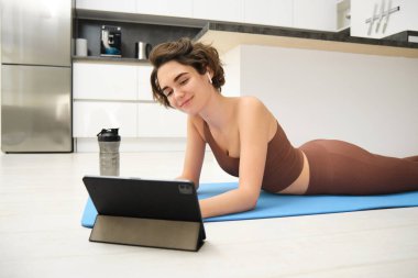Tüm vücuduyla gülümseyen genç, güçlü spor spor spor spor antrenörü kadın ev içinde spor salonunda egzersiz yapmak için kahverengi eşofman giyecek ve tablet kullanacak. Spor sporu konsepti.