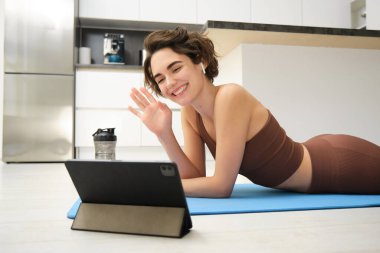 Gülümseyen esmer kadın spor giyim, el sallıyor, tabletten selam veriyor, video spor blogunu kaydediyor, fitness antrenmanını internetten yapıyor, eğitmen egzersiz unsurlarını gösteriyor, mutfakta lastik paspas kullanıyor..