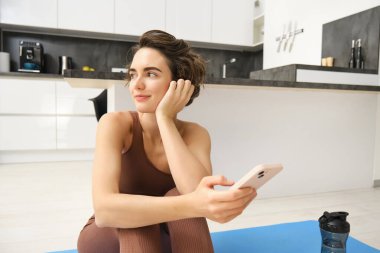 Cep telefonlu sportif bir kızın portresi, yoga minderinde oturmak, antreman sırasında akıllı telefon kullanmak, telefonda spor salonunu izlemek. Spor ve refah kavramı