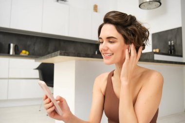 Güzel, gülümseyen fitness kadınının portresi, akıllı telefona bakıyor, video görüşmeleri fitness eğitmeni, evden online beden eğitimi dersi alıyor, telefona bakıyor, kablosuz kulaklık takıyor, uzaktan antrenman yapıyor..