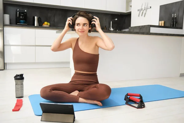 Gym at home. Young smiling woman puts on headphones in her kitchen, wearing sportswear, ready for workout, follows fitness instructor app online video on tablet, sits with exercise equipment.
