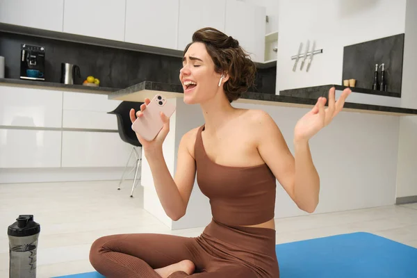 stock image Carefree fitness girl, workout instructor sits at home with water bottle, yoga mat, wears tracksuit, sings into her smartphone, enjoys music in wireless headphones, listens song during pilates yoga.