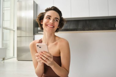 Aktif giyimli spor bir kadın görüntüsü, evde yoga minderinde oturuyor, akıllı telefonu tutuyor, egzersiz uygulaması kullanıyor, çevrimiçi egzersiz arıyor. Boşluğu kopyala