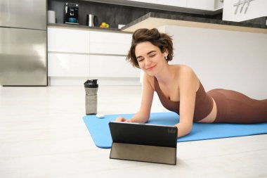 Genç fitness kadını, eşofmanlı kız fitness minderinde yatıyor, yogaya bakıyor, tablete pilates video dersi veriyor, evden antrenman yapıyor, spor uygulaması kullanıyor, evde eğitim görüyor..