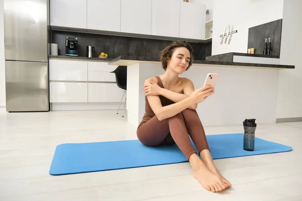 Portrait of sporty girl with mobile phone, sitting on yoga mat, using smartphone during workout at home, watching fitness turorial on telephone. Sport and wellbeing concept