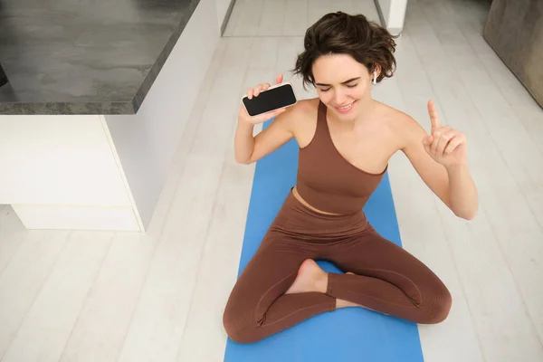 Top view of young active woman, healthy girl stretching, doing workout at home, holding smartphone and listening music in headphones while workout at home, does fitness, pilates yoga on floor mat.