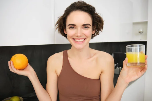 Smiling sportswoman showing an orange and glass of juice, sharing tips for healthy and active lifestyle, standing at home in kitchen.