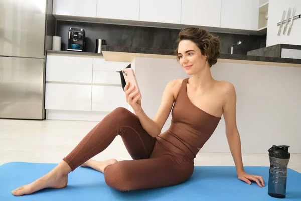 stock image Young fitness instructor looking at her smartphone, sitting on rubber mat at home. Young woman doing workout training, looking at exercise videos on mobile app.