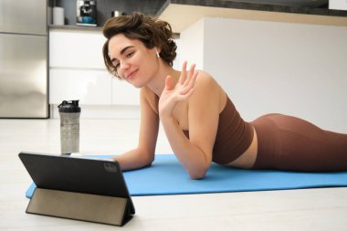 Spor ve yaşam tarzı. Genç fitness kadını, beden eğitimi hocası tablette el sallıyor, çevrimiçi video sınıfına katılıyor, yoga ve farkındalık egzersizi yapıyor, evde antrenman yapıyor..