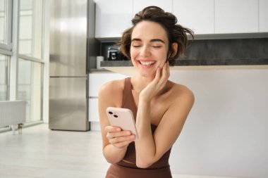 Aktif giyimli spor bir kadın görüntüsü, evde yoga minderinde oturuyor, akıllı telefonu tutuyor, egzersiz uygulaması kullanıyor, çevrimiçi egzersiz arıyor. Boşluğu kopyala