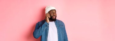 Hipster siyahi adam telefonda konuşuyor, sola bakıyor ve telefonla konuşuyor, pembe arka planda duruyor..