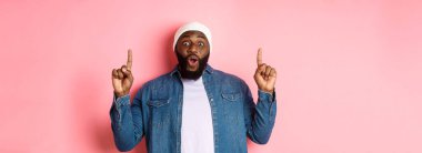 Heyecanlı ve etkilenmiş siyah adam harika bir tanıtım sergiliyor, parmaklarını logoya doğrultuyor, pembe arka planda duruyor..