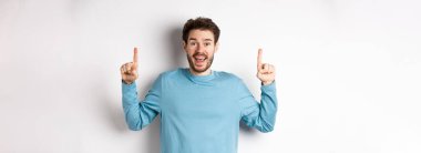 Neşeli genç adam mutlu bir gülümsemeyle reklamını yapıyor, parmaklarını muhteşem logo afişini işaret ediyor, beyaz arka planda duruyor..