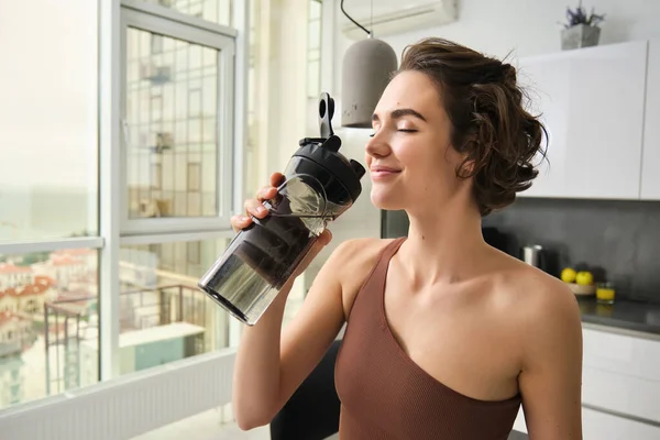 Sport and fitness concept. Portrait of young woman athlete drinking water from gym bottle, smiling with pleased face, wearing acrivewear, standing in kitchen.