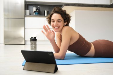 Spor ve yaşam tarzı. Genç fitness kadını, beden eğitimi hocası tablette el sallıyor, çevrimiçi video sınıfına katılıyor, yoga ve farkındalık egzersizi yapıyor, evde antrenman yapıyor..