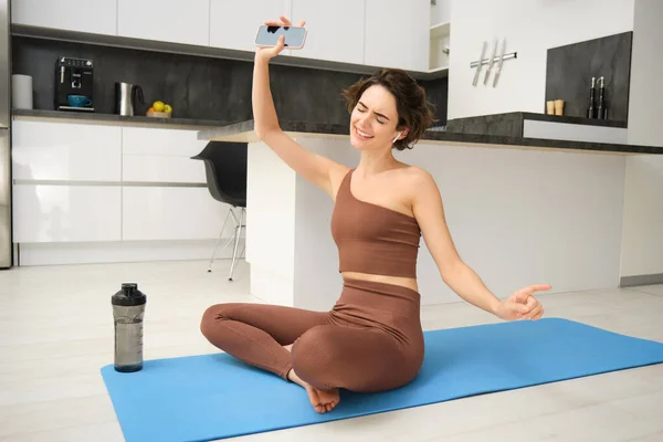 stock image Happy brunette woman, sporty girl in activewear, dancing while sitting on yoga mat, listens music during sport workout at home.