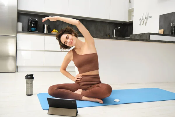 Young woman looking at digital tablet, connects to stretching fitness online class, workout, sits on yoga rubber mat at home, follows gym instructor exercises remotely.