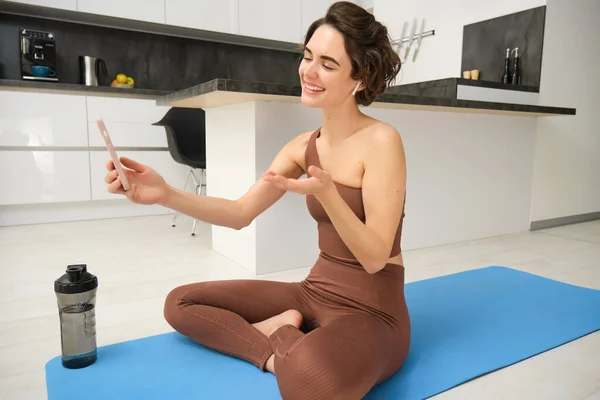 stock image Portrait of happy smiling, relaxed fitness instructor, girl video chats, looks at smartphone, records workout blog, yoga online, sits on sports mat in kitchen, does pilates from home.