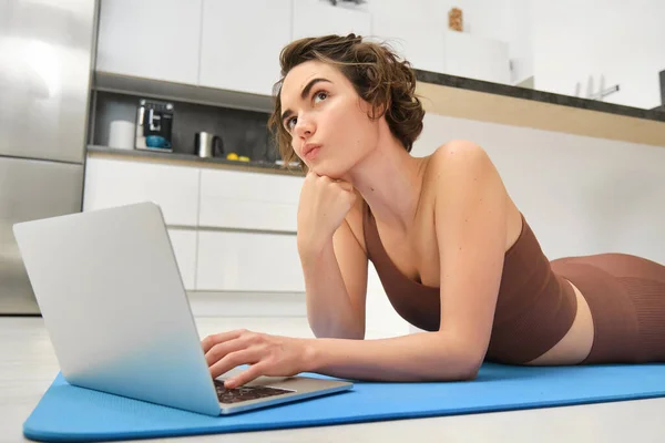 年轻女运动员的画像 躺在地板上 用瑜伽垫 一边用笔记本电脑思考 在室内找体操练习 体育和健身概念 — 图库照片