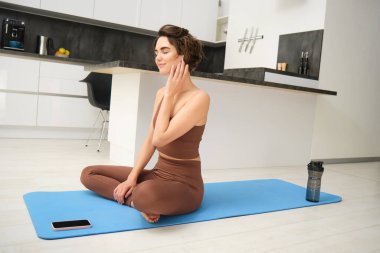 Farkındalık ve sağlıklı yaşam tarzı. Genç, gülümseyen bir kadın evde oturmuş, kulaklıkla yoga yapıyor, akıllı telefonda meditasyon uygulaması dinliyor, egzersiz ve egzersiz için paspas kullanıyor..
