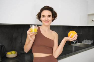 Sağlıklı yaşam tarzı ve spor. Güzel, gülümseyen bir kadın, taze portakal suyu içiyor ve elinde meyve tutuyor, evde egzersiz yaptıktan sonra vitamin içeceğinin tadını çıkarıyor, mutfakta duruyor..