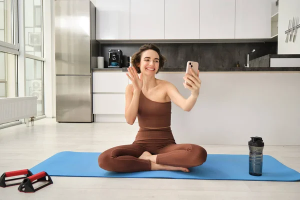 Image of young fitness girl blogger, records her workout at home, chats online while doing yoga in bright room. Woman with smartphone, joining remote yoga training via mobile phone.